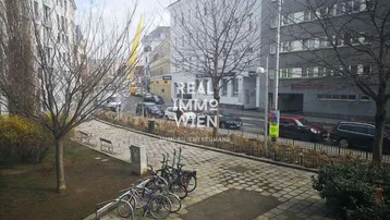 Blick auf eine belebte Stadtstraße mit parkenden Autos, Bäumen und einem kleinen Grünstreifen mit Fahrradständern.