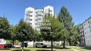 Mehrstöckiges Wohngebäude mit Balkonen und grüner Umgebung unter blauem Himmel.