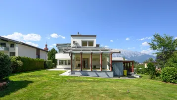 Modernes Haus mit großem Garten, Wintergarten und Bergblick unter blauem Himmel.