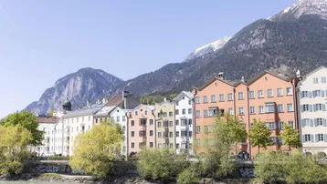 Blick auf die bunte Häuserzeile entlang des Flusses mit schneebedeckten Bergen im Hintergrund.