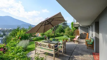 Große Terrasse mit Holzboden, Gartenmöbeln und einem Sonnenschirm, umgeben von üppigem Grün und Bergblick.