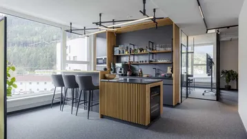 Moderne Büroküche mit Holzakzenten, Barhockern und großen Fenstern mit Bergblick.