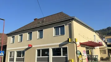 Gebäudeansicht von der Straßenseite mit hellbeiger Fassade und großen Fenstern im Erdgeschoss, unter blauem Himmel.