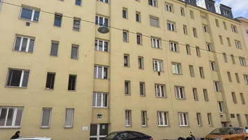 Ansicht der gelben Fassade eines Mehrfamilienhauses mit vielen Fenstern und geparkten Autos auf der Straße.
