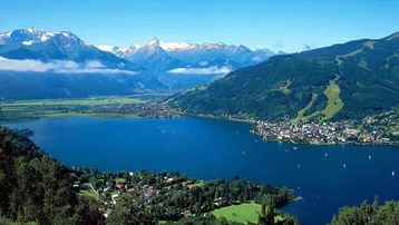 Panoramablick auf einen malerischen See, umgeben von grünen Hügeln, einer Stadt und schneebedeckten Bergen.