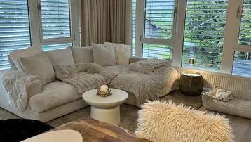 Gemütliches Wohnzimmer mit großem Sofa, hellen Vorhängen und weitem Blick ins Grüne durch große Fenster.