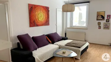 Gemütliches Wohnzimmer mit Sofa, Couchtisch und farbenfrohem Wandbild, hell durch großes Fenster.