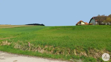 Ländliche Umgebung mit weiten Grünflächen und Häusern im Hintergrund unter blauem Himmel.