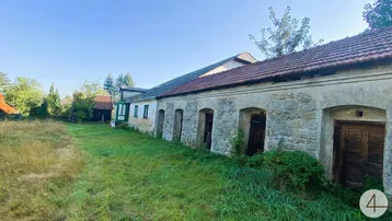 Lange Fassade eines alten Gebäudes mit Steinbögen und einer weiten, ungemähten Wiese davor unter blauem Himmel.