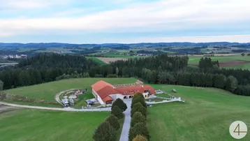 Luftaufnahme eines weitläufigen Anwesens mit rotem Dach, umgeben von grünen Hügeln und Wäldern unter bewölktem Himmel.