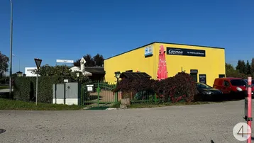 Gelbes Geschäftsgebäude mit roten Akzenten, umgeben von Hecken und Parkplätzen unter blauem Himmel.
