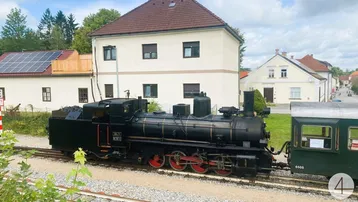 Historische Dampflokomotive auf Gleisen vor ländlichen Gebäuden mit Solaranlage auf dem Dach.