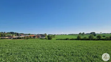 Weitläufige grüne Landschaft mit Feldern und Häusern in der Ferne unter klarem blauem Himmel.
