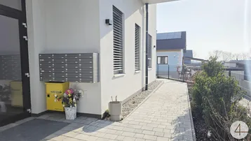 Eingangsbereich eines modernen Mehrfamilienhauses mit Briefkästen und gepflastertem Gehweg unter blauem Himmel.