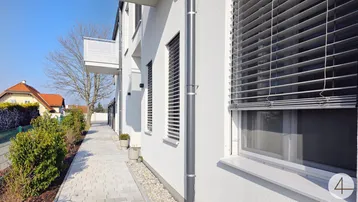 Moderne Gebäudefassade mit weißen Balkonen, Jalousien und gepflastertem Weg an einem sonnigen Tag.