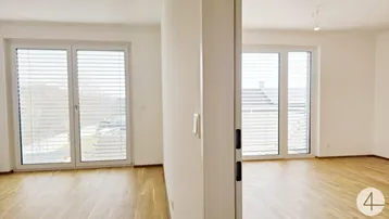 Helles, leeres Zimmer mit Parkettboden und großen Fenstern, die viel Tageslicht hereinlassen.