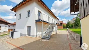 Modernes weißes Gebäude mit externer Metalltreppe, die zu einem Balkon führt, und gepflastertem Vorplatz.