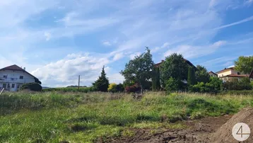 Ungenutztes Grundstück mit Grasfläche und Häusern im Hintergrund unter blauem Himmel.