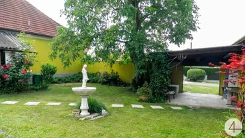 Grüner Garten mit Rasenfläche, Bäumen, einem dekorativen Brunnen und einem gelben Haus im Hintergrund.