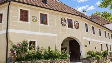 Historisches Gebäude mit beiger Fassade, Rundbogenportal und traditionellen Fensterläden, umgeben von Rosensträuchern.