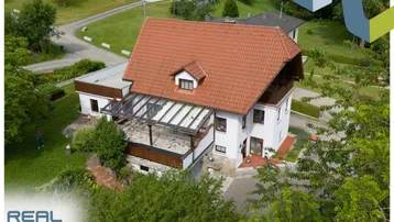 Luftaufnahme des Einfamilienhauses mit rotem Ziegeldach, Terrasse und umgebender grüner Landschaft.