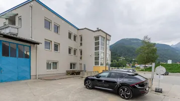 Gebäudeansicht mit moderner Architektur, großen Fenstern und einem geparkten Elektrofahrzeug vor einer Bergkulisse.