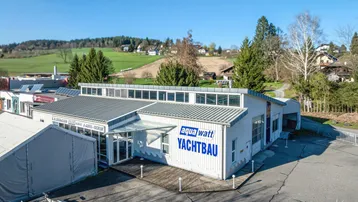 Luftaufnahme des Gebäudes mit dem Schriftzug 'aqua watt YACHTBAU' und umliegender grüner Landschaft unter blauem Himmel.