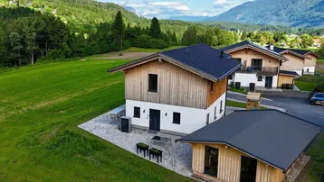 Moderne Häuser im alpinen Stil mit Holzfassaden, umgeben von grünen Wiesen und Bergen.