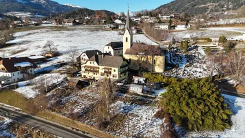 Luftaufnahme eines verschneiten Dorfes mit Kirche und Bergen im Hintergrund unter blauem Himmel.