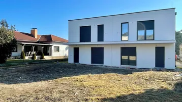 Modernes, weißes Neubauhaus mit dunklen Fensterläden und unbebautem Vorgarten.