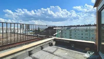 Unfertige Dachterrasse mit Metallgeländer und Blick auf die umliegende Stadtlandschaft unter blauem Himmel.