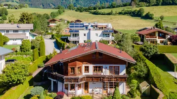 Traditionelles Haus mit Holzelementen und rotem Dach, umgeben von grünen Hügeln und Bergen.