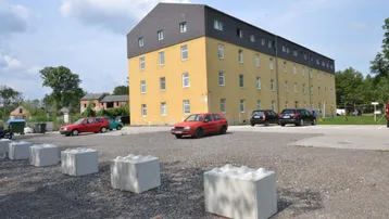 Gelbes Mehrfamilienhaus mit dunklem Dach und großem Schotterparkplatz davor unter bewölktem Himmel.