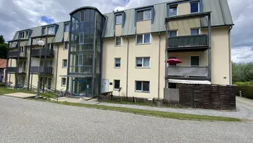 Mehrfamilienhaus mit moderner Glasfassade am Eingang und mehreren Balkonen unter bewölktem Himmel.