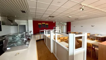 Innenansicht eines Restaurants oder einer Kantine mit Thekenbereich, Tischen und Stühlen sowie roter Akzentwand.