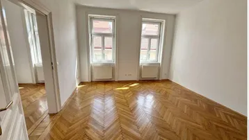 Heller Wohnraum mit Fischgrätparkettboden, weißen Wänden und zwei Fenstern, die viel Tageslicht hereinlassen.