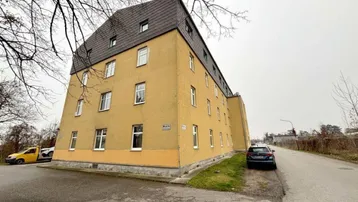 Mehrstöckiges Gebäude mit gelber Fassade und Mansarddach an einem bewölkten Tag.