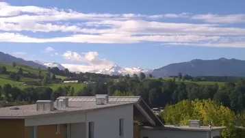 Bergblick über die Dächer von Gebäuden mit grünen Hügeln und schneebedeckten Gipfeln im Hintergrund.