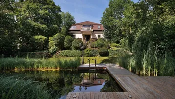 Elegante Villa mit weitläufigem Garten, Teich und Holzsteg unter blauem Himmel.