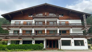 Traditionelles Gebäude mit Holzfassade und mehreren Balkonen, umgeben von grüner Landschaft und Bergen.