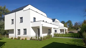 Moderne weiße Fassade eines Neubaus mit klaren Linien und gepflegtem Rasenbereich unter blauem Himmel.