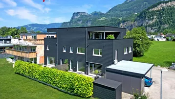 Modernes Mehrfamilienhaus mit dunkler Fassade, umgeben von grünen Wiesen und Bergen im Hintergrund.