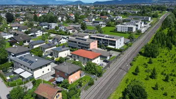 Weitläufige Luftaufnahme der Wohnsiedlung mit Bahngleisen und Bergpanorama im Hintergrund, sehr idyllisch.