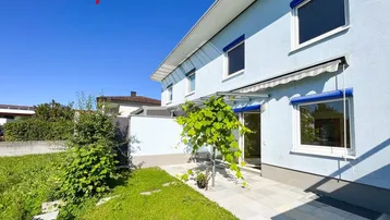 Seitlicher Blick auf das Haus mit einer kleinen gepflasterten Terrasse und einem grünen Gartenbereich.
