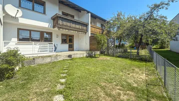 Hausrückseite mit Garten, Terrasse und Balkon unter blauem Himmel.