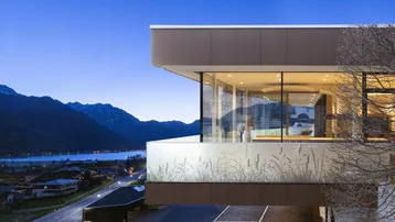 Moderne Architektur mit großen Glasfronten und Panoramablick auf See und Berge bei Dämmerung.