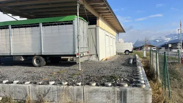 Außenansicht eines modularen Gebäudes mit Holzdach und einem Anhänger auf einem Schotterplatz vor Bergkulisse.