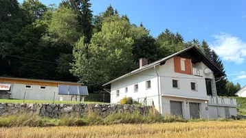 Ansicht des Hauses und eines Nebengebäudes mit Solaranlage, umgeben von Feldern und Wald.