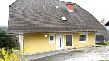 Gelbes Einfamilienhaus mit dunklem Schindeldach, umgeben von Grün und einem kleinen Gartenbereich.