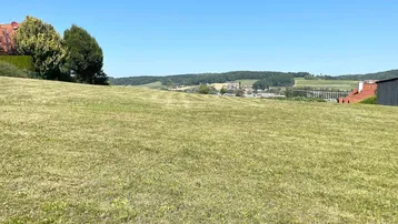 Weitläufige grüne Wiese mit Blick auf die umliegende Landschaft und entfernte Häuser unter blauem Himmel.